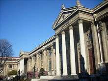 The façade of a masonry building, with four Greek adorning its entrance, under a clear blue sky