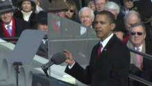 Barack Obama in a black coat gestures and talks at a lectern surrounded by teleprompters. Behind him, in the background, are about a dozen warmly dressed onlookers.