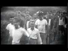 Video of men walking in a line, who look at and smile at the camera. They are led by a man riding a carabao pulling a cart. The men wear assorted vests, T-shirts, uniforms, shorts, and pants. Brush can be seen in the background.