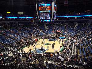 Scattered persons are present in a blue seating area surrounding a basketball court with several players warming up. A large scoreboard with video monitor is visible above the court.