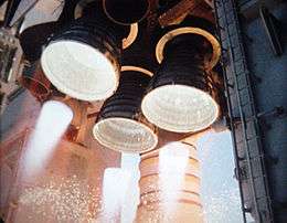 Three bell-shaped rocket engine nozzles projecting from the aft structure of a Space Shuttle orbiter. The cluster is arranged triangularly, with one engine at the top and two below, with two smaller nozzles visible to the left and right of the top engine. The three larger engines are firing, with white-hot flames visible projecting from each nozzle. The Space Shuttle's left Solid Rocket Booster (a white, cylindrical rocket) is visible in the background, with the two large, grey tail service masts visible to the left and right of the orbiter's aft structure.