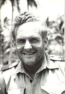 Informal head-and-shoulders portrait of grinning man in light-coloured shirt with palm trees in background