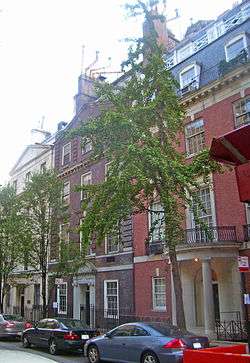 Three townhouses with two trees in front and ornate facades seen from across the street and to their right. They are all three stories high. The one on the left is light tan, the center one dark red brick and the left one regular red brick.