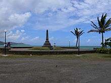 A stretch of a bay. In the foreground is a gravel area in front of a large obelisk and two palm trees.