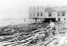 A black and white image of a large, scattered pile of wooden planks upon which a man in a white shirt is walking. In the background a building with windows and stilts is visible.