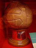 12-panel brown leather football on a plinth of hardwood, with a brass panel inscribed with the names of the victorious team members, photographed through the glass of a display case