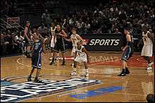 Two opposing basketball teams on a basketball court with a team in white defending the basket behind them. One team is in white uniforms with the word Michigan on the front and names on the back and the other is dark blue with the word Duke on the front and names on the back.