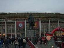 A wide shot of a large, bronze statue of Lenin on a tall plinth. In the background is a stadium draped with red banners, one with the Manchester United crest and one with the Chelsea crest. People are walking towards the stadium and a Russian policeman is in the foreground.