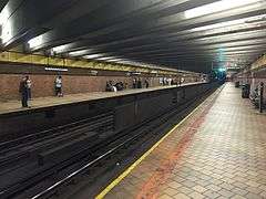 People waiting on a dimly lighted, nearly empty brick-tile platform at the 21st Street – Queensbridge station