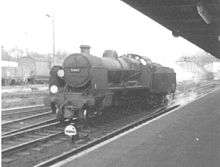 Side-and-front view of the rebuilt form of the locomotive, running without a train through a railway station. The locomotive is of conventional appearance, with a tender and has no side watertanks obscuring the boiler. Smoke deflectors are fitted at the front of the locomotive.