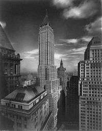 Distant aerial view of a 70-story building with several setbacks and a pyramidal roof; a flagpole sits stop the roof.