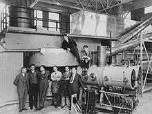 Six men in suits and ties stand stand in front of gigantic machinery. Two more are sitting in top of it.