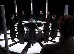 A group of men and women stand in a circle, they are all wearing black robes.