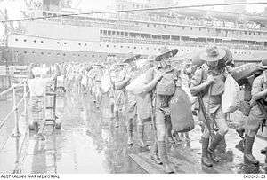 Men in shorts and slouch hats with rifles slung, carrying duffel bags march along a wharf. In the background is a cruise ship.