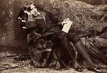 A tall man rests on a chaise longue, facing the camera. On his knees, which are held together, he holds a slim, richly bound book. He wears knee breeches which feature prominently in the photograph's foreground.