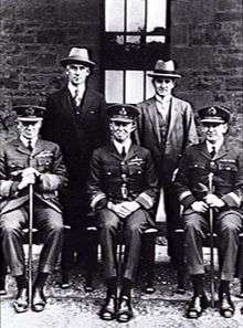 Formal portrait of three men in military uniforms with peaked caps sitting in front of two men in civilian clothes with broad-brimmed hats