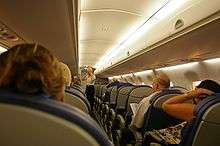 Interior of single-aisle jet aircraft, with people seated. Light illuminate the ceiling as a female stands near the front of aircraft