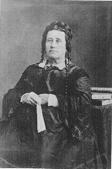 Portrait of an unsmiling, middle-aged woman in a voluminous dress. Her hair is piled on the back of her head, with ringlets near her ears. She holds a fan in her hands.