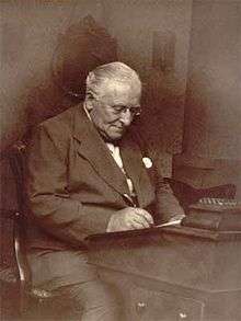 Photograph of a man, with white hair and wearing a suit, sitting working at a desk