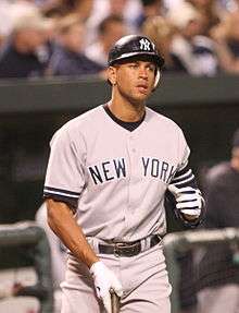 A man in a dark batting helmet, grey baseball uniform, and white gloves holds a baseball bat against the ground with his right hand.