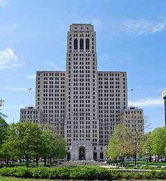 A tall white blocky stone building seen from across a square with trees and a walkway leading to the arched main entrance. Just below the top are three similar long narrow arched windows; the building has two wings on either side, about two-thirds its height, which themselves have wings about half their height from which American flags fly on poles on top.