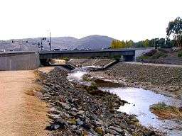 A channelized stream between rocky banks runs towards a canyon in the distance as it flows under a concrete bridge