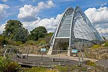 A narrow semicircular building of glass and steel latticework stands at the right, set amid an area of worked rock with a line of deciduous trees in the rear left, under a blue sky filled with large puffy white clouds. In front of it, curving slightly away to the left, is a wooden platform with benches on it and a thin metal guardrail in front of a low wet area with bright red flowers