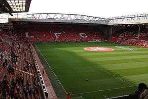 A two-tiered stand which has red seats, there are also white seats which spell out "L.F.C.. In front of the stand is a field of grass