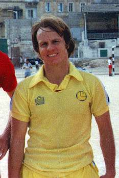 An upper-body photograph of a man with medium-length brown hair wearing a yellow shirt.