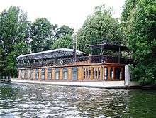 A colour image Gilmour's houseboat and studio the Astoria, anchored in a river. The background is green forest and it is a bright sunny day.