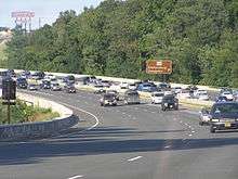 A tree lined six lane freeway with congestion on the right side of the road surrounded by trees. To the right, a brown sign reads Maryland Route 450 Bladensburg Annapolis with an arrow pointing to the upper right. A sign for the Capital Plaza Mall is in the distance