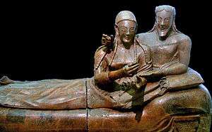 A sculpture of a woman and man reclining together on a couch, their upper bodies to the right and their legs to the left. There is a marked contrast between the high relief busts of their upper bodies and the very flattened lower bodies and legs. They have almond-shaped eyes and long braided hair, and are smiling widely. The man has a beard that is bobbed. The woman's hands are gesticulating in front of her as if she was holding something that is no longer there, or perhaps gesturing while speaking. The man has his right arm draped around the woman's shoulders in an intimate pose, and his right hand on her shoulder also appears to once have held some item. His left hand rests palm up in the crook of the woman's left elbow.