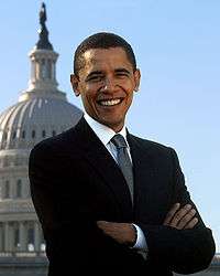 An African-American man in a black suit, a grey tie, and the U.S. Capitol dome behind the subject in the distance.