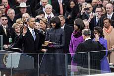 Barack Obama holds his right hand in the air as Michelle Obama looks at him and Malia and Sasha Obama watch as a man whose back is to the camera looks down.