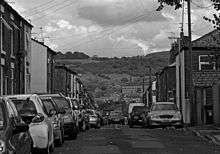 A black and white view of a street with parked cars and terraced brick houses on both sides gently sloping downhill, revealing a view of a hillside with a large brick industrial building on it