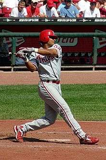 A man in a gray baseball uniform with red trim swinging a black baseball bat