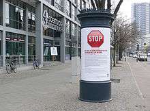 A sidewalk, bordered on the left by a modern building featuring the inscription "Scientology-Kirche" above its entrance and on the right by a cycle path, beyond which there is a road with a yellow "H" sign marking a bus stop. The pavement is lined by trees on the side facing the cycle path. In the foreground there is an advertising column with a white poster featuring a large red Stop sign, followed by text commenting on the activities of Scientology in the local area. A corner of the roof of a bus shelter is visible protruding behind the column.