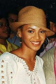 An image of a smiling woman wearing large hoop earrings and a beige hat. She is also wearing a white blouse and people can be seen standing behind her.