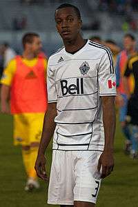 A man wearing a white t-shirt and white shorts, standing on a soccer field with his hands at his side, looking ahead. In the background, a man wearing a yellow t-shirt with an orange bib over top, yellow shorts, and yellow socks.