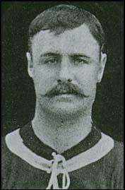 A black-and-white head-and-shoulders photograph of a man wearing a dark shirt with a light ring around the collar. He has short hair and a moustache.