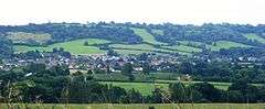 The roofs of many houses can be seen in a green valley with several trees.