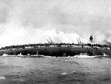 The burning hull of a ship on its side; water pours from holes punched by enemy projectiles. The keel is splitting from the intense strain.