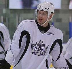 Head and upper body of a hockey player in white uniform with a picture of a crown in the middle. He smiles slightly, looking forward and to his right.