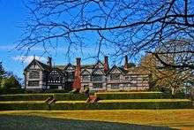 Exterior view of the side of a large black and white building with numerous chimneys. In the foreground are two rows of hedges.