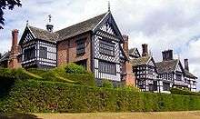 The exterior of a large house. There are several chimneys, leaded windows and wings. In the foreground are two rows of hedges.