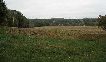 Photo from a hill top showing trees on the left and a plowed field sloping down. In the distance, the land slopes upward among trees.