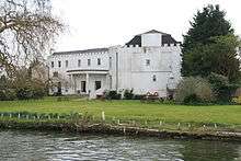 Colour photograph of a large white building with a castellated roof surrounded by trees situated in front of a river