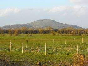 Large green hill, partially covered in pasture and trees, with more trees, meadows, and wire fences on the level ground in the foreground of the picture