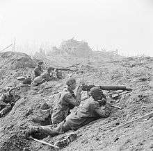 Five men with two guns in a devastated landscape. The remains of a bombed town are in the background