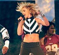 A blonde female performer in a black-and-white ensemble, holding a microphone near her mouth